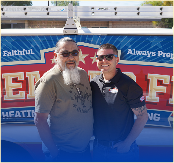 Two men smiling and posing in front of a blue and red promotional van with a banner outdoors on a sunny day.