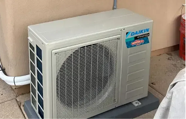 Outdoor Daikin air conditioner condenser unit mounted on a concrete pad beside a wall, with a visible fan grille and labeled Daikin component.
