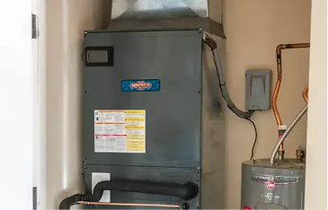 Gray furnace in a utility room beside a vertical water heater, with exposed copper pipes and wiring on the wall below a small electrical box.