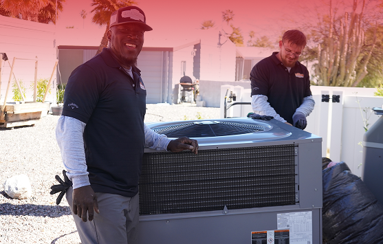 Two HVAC technicians in dark polo shirts work on a large outdoor air conditioning condenser in a sunny yard, smiling at the camera.