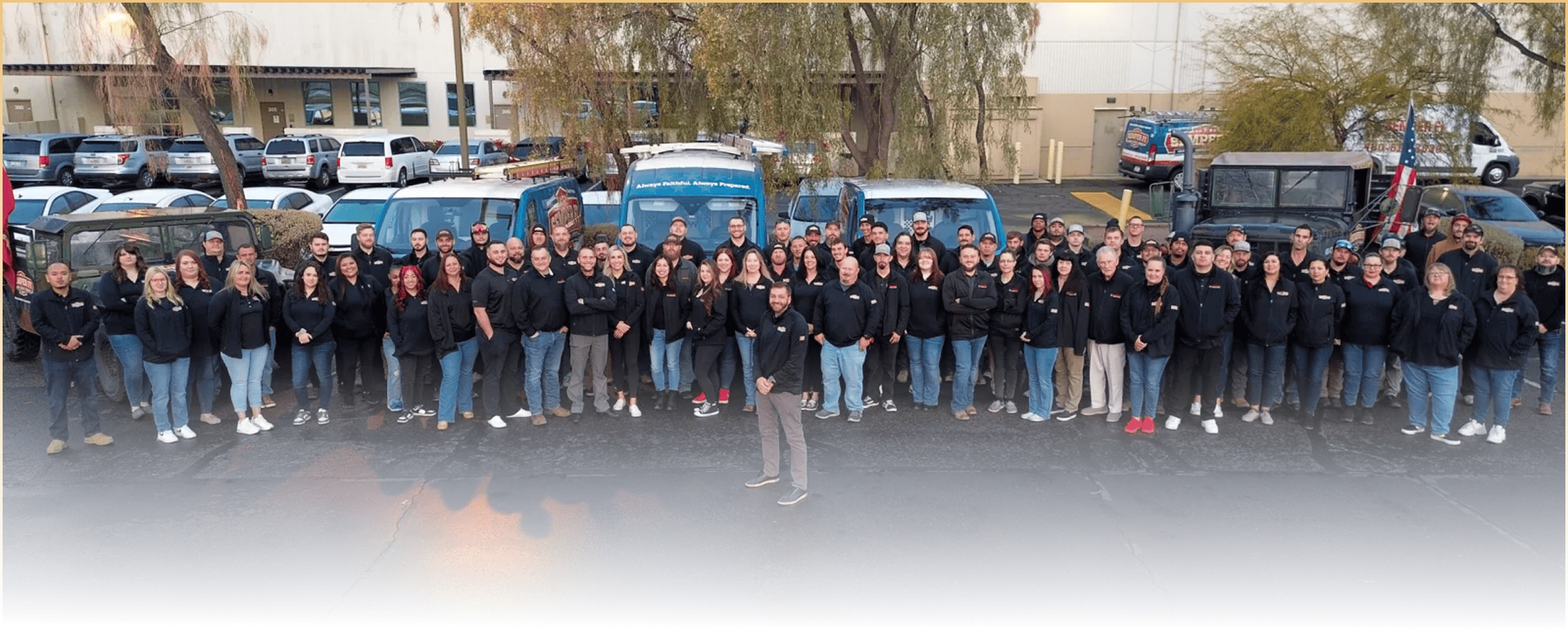 Group of employees in black jackets posing outside in front of service vans and trucks for a company photo.