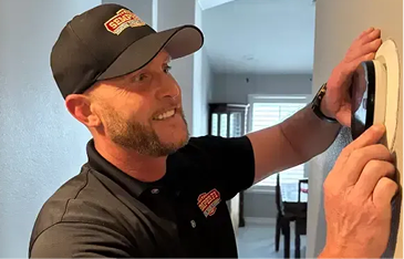 Home repair technician in a black polo and cap installs a round, wall-mounted device, smiling as he presses it into place in a home interior.