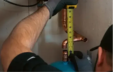 Worker wearing a glove measures a copper pipe with a yellow tape measure under a sink, preparing a cut or fitting.