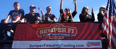Group of people on a parade float holding a Semper Fi Heating Cooling Plumbing banner with an American flag in the background.