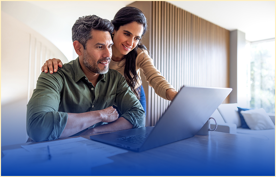 Couple sits at a laptop on a coffee table, smiling as they work together at home.