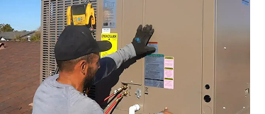 HVAC technician wearing a cap and gloves inspecting an outdoor air conditioner/heat pump unit on a sunny rooftop.
