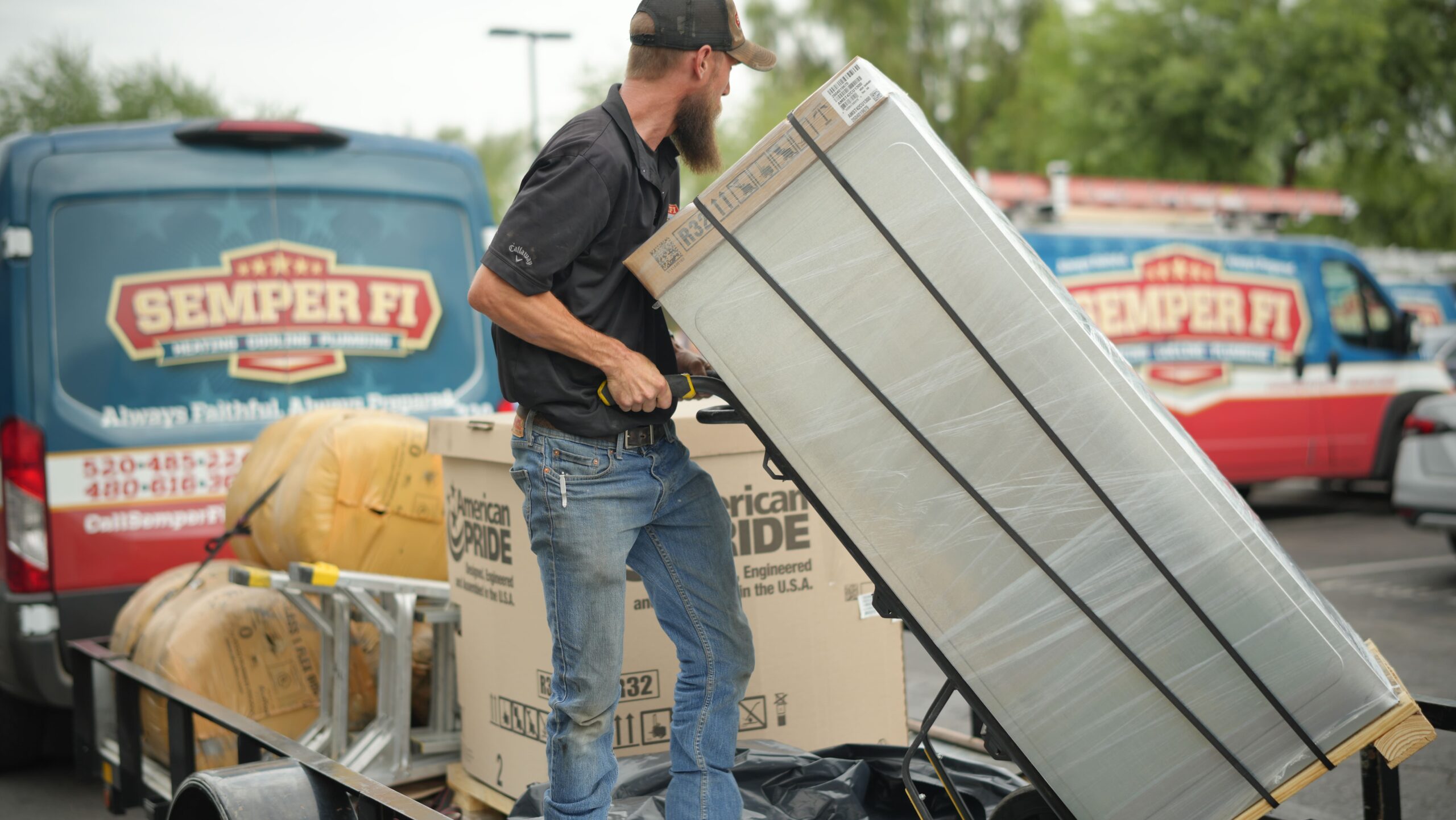 Volunteer with a beard and cap lifting a large metal appliance onto a trailer during a charity donation event, with donation boxes and a Semper Fi van in the background.